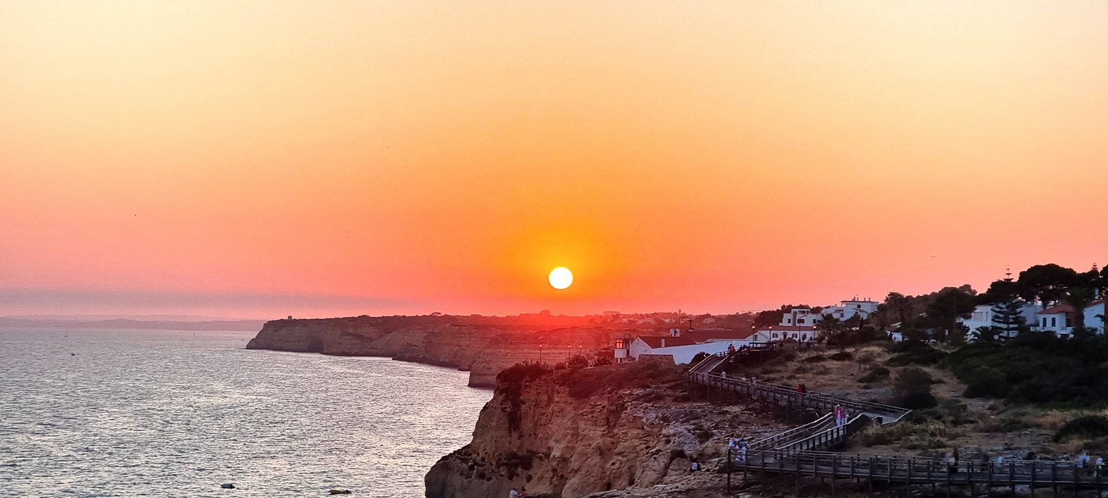Ciel orange, le soleil bas au-dessus de la côte ; des promeneurs sur le ponton et les maisons blanches du village au bout de la falaise